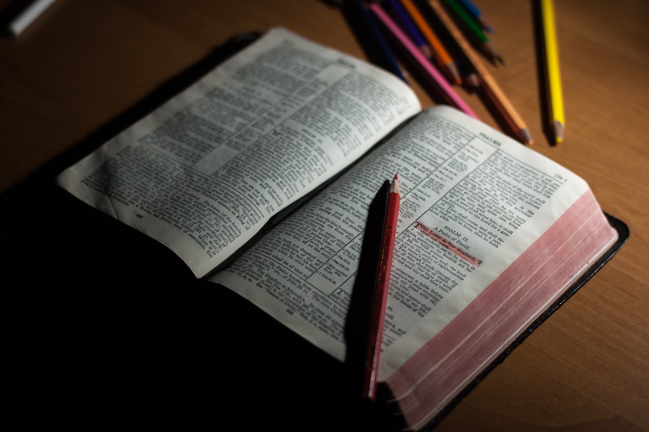Contact US An open Bible on a wooden table accompanied by colored pencils, symbolizing study and spirituality.